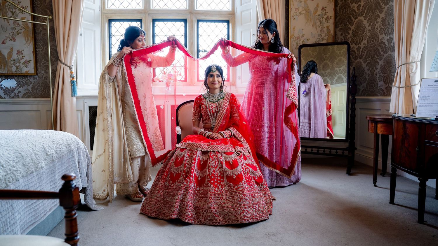 Indian bride in red lehenga seated as bridesmaids hold her dupatta in an elegant room.