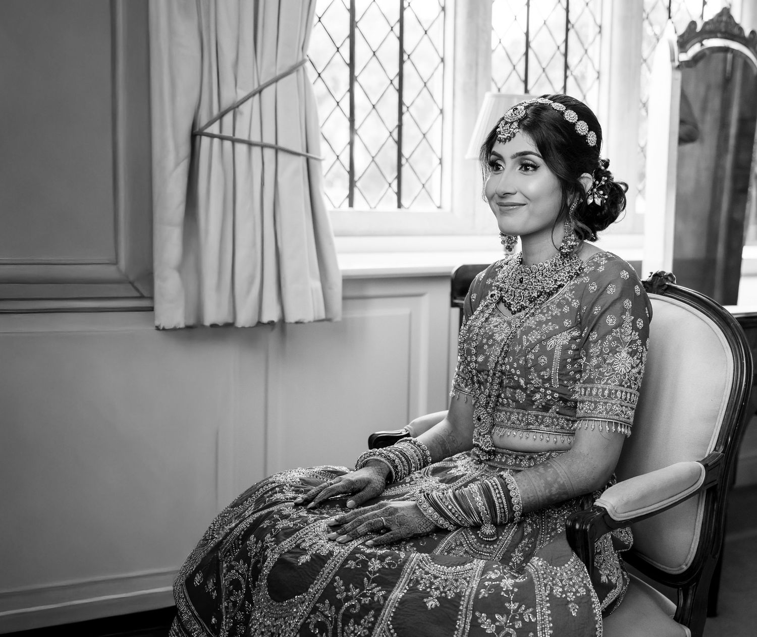 Black and white portrait of Indian bride in embroidered lehenga seated by window wearing bridal jewelry.