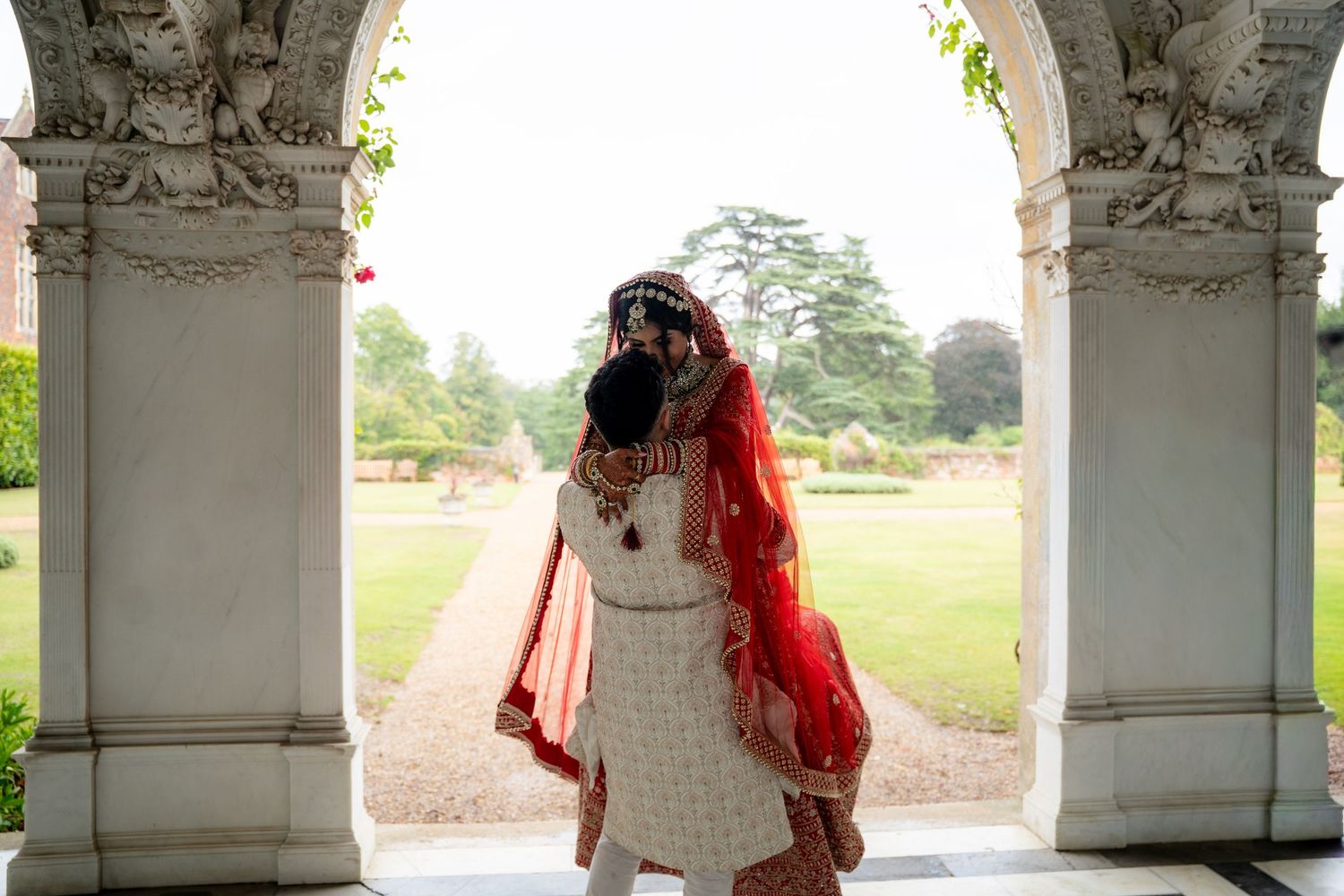Indian bride in red and white lehenga standing under ornate white stone archway in lush garden setting.