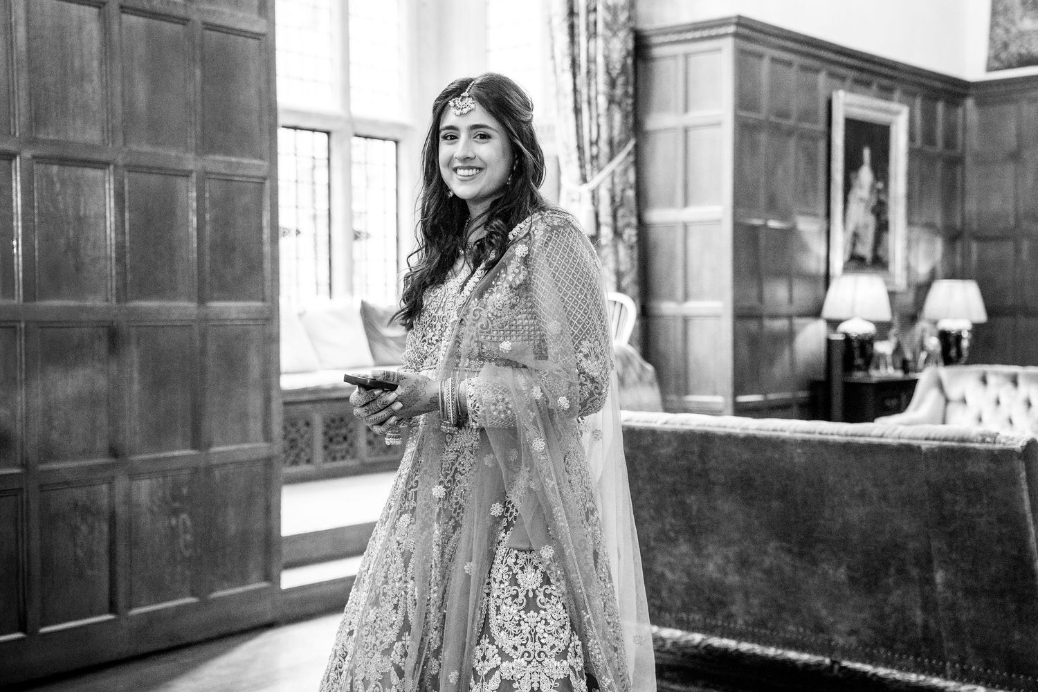 Black and white photo of smiling woman in embroidered lehenga holding a phone in ornate room.