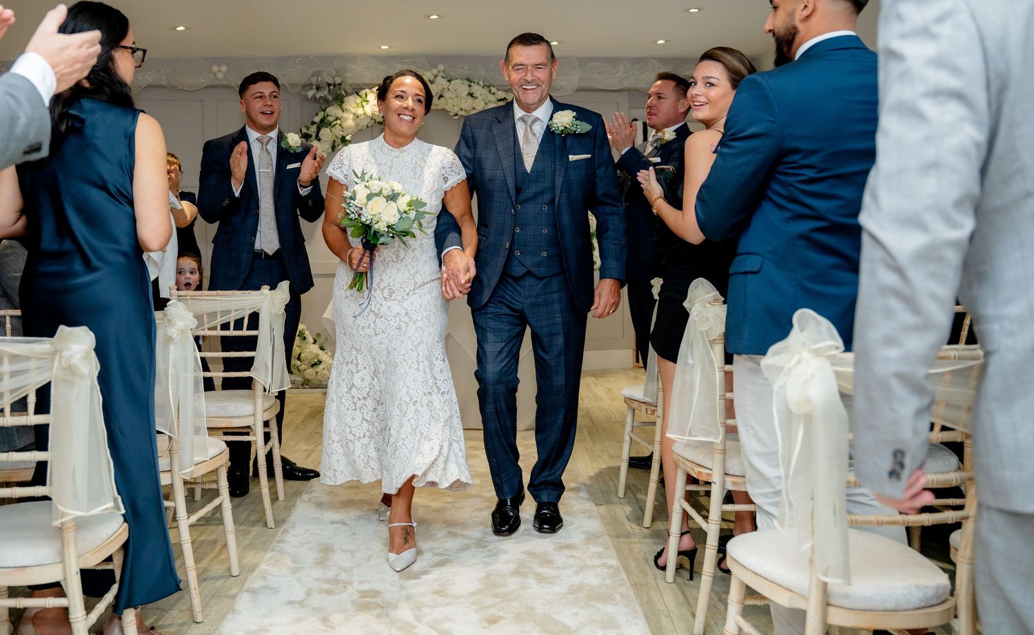 Newlyweds walking down aisle holding hands while guests celebrate in bright wedding venue.