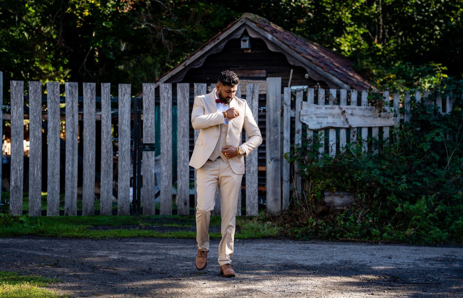 Groom in cream suit adjusts jacket while walking on a sunny garden path.