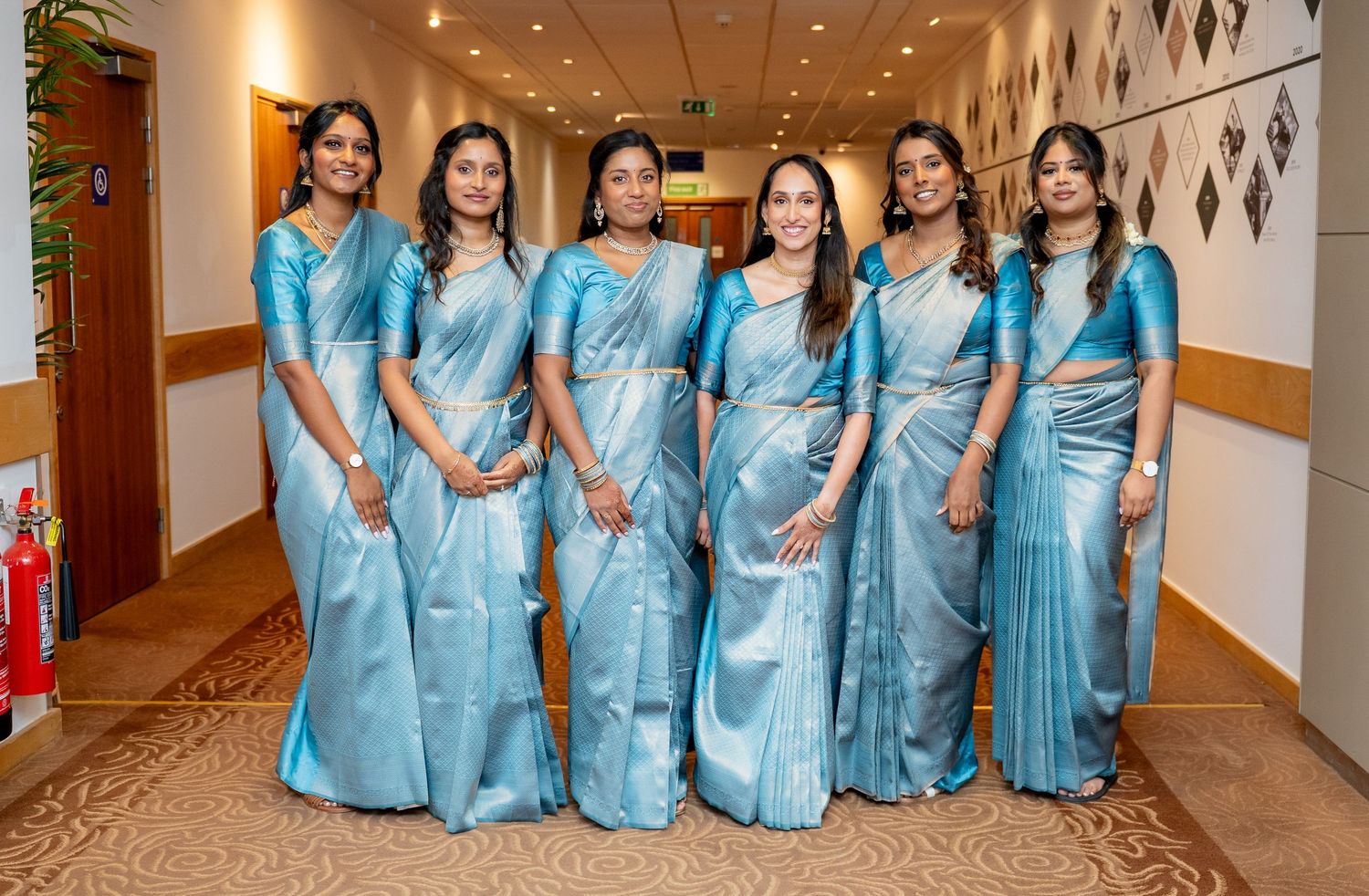 Six women posing together in matching blue silk sarees at a formal Indian wedding celebration.