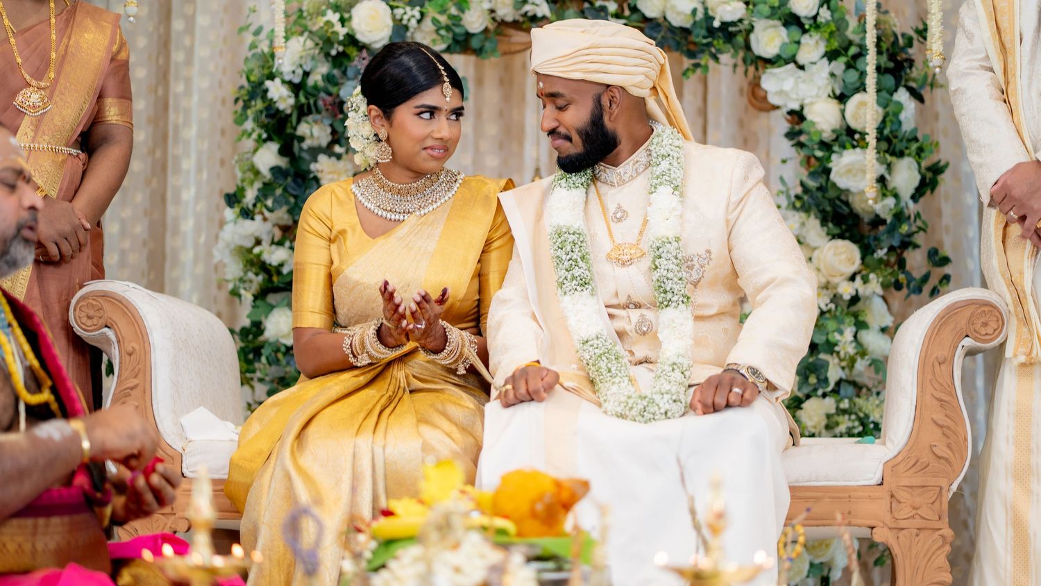 Indian bride in gold silk saree and groom in cream sherwani with floral garlands at Hindu wedding ceremony.