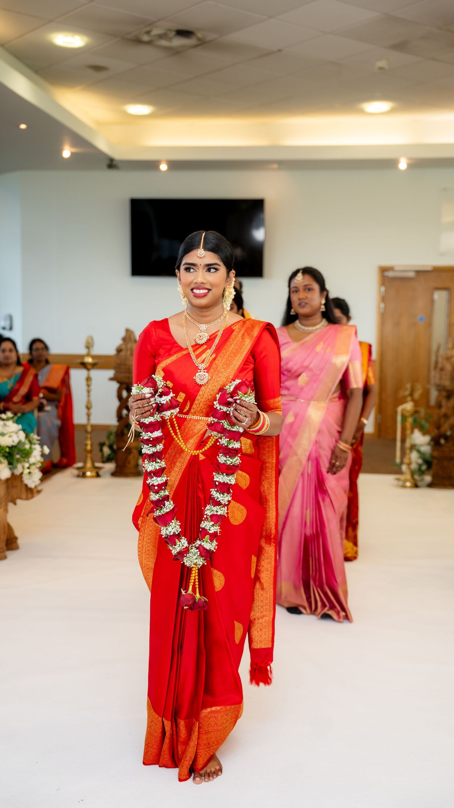 South Indian bride in red silk saree walks down decorated wedding hall aisle with garlands.