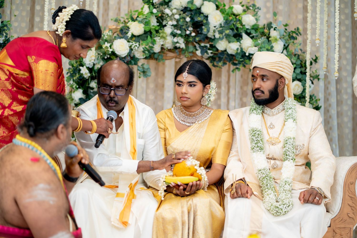 South Indian wedding ritual with elderly man speaking at microphone while bride holds ceremonial bowl at altar.