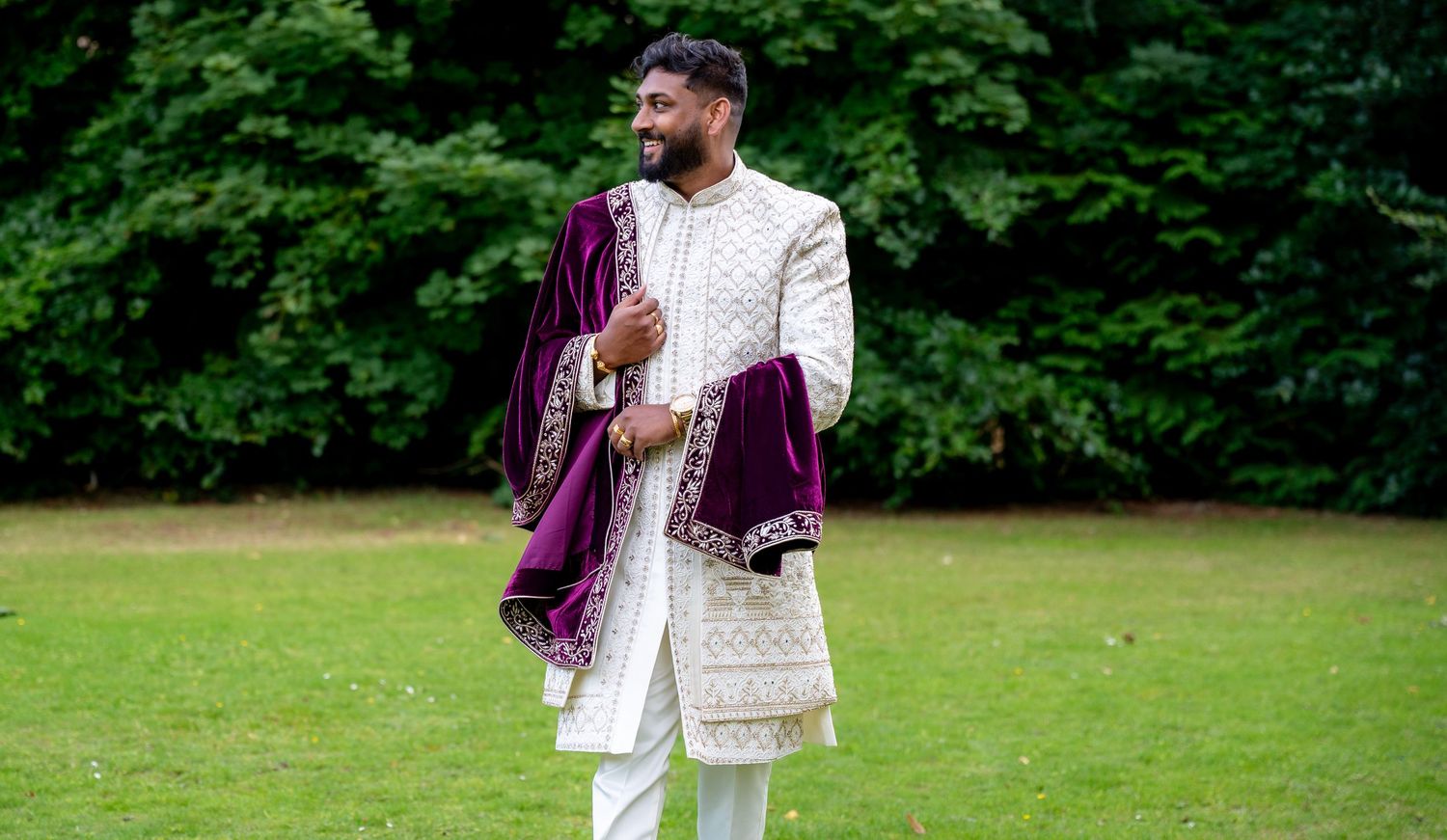 Smiling groom in white embroidered sherwani with burgundy velvet dupatta posing in lush green garden.