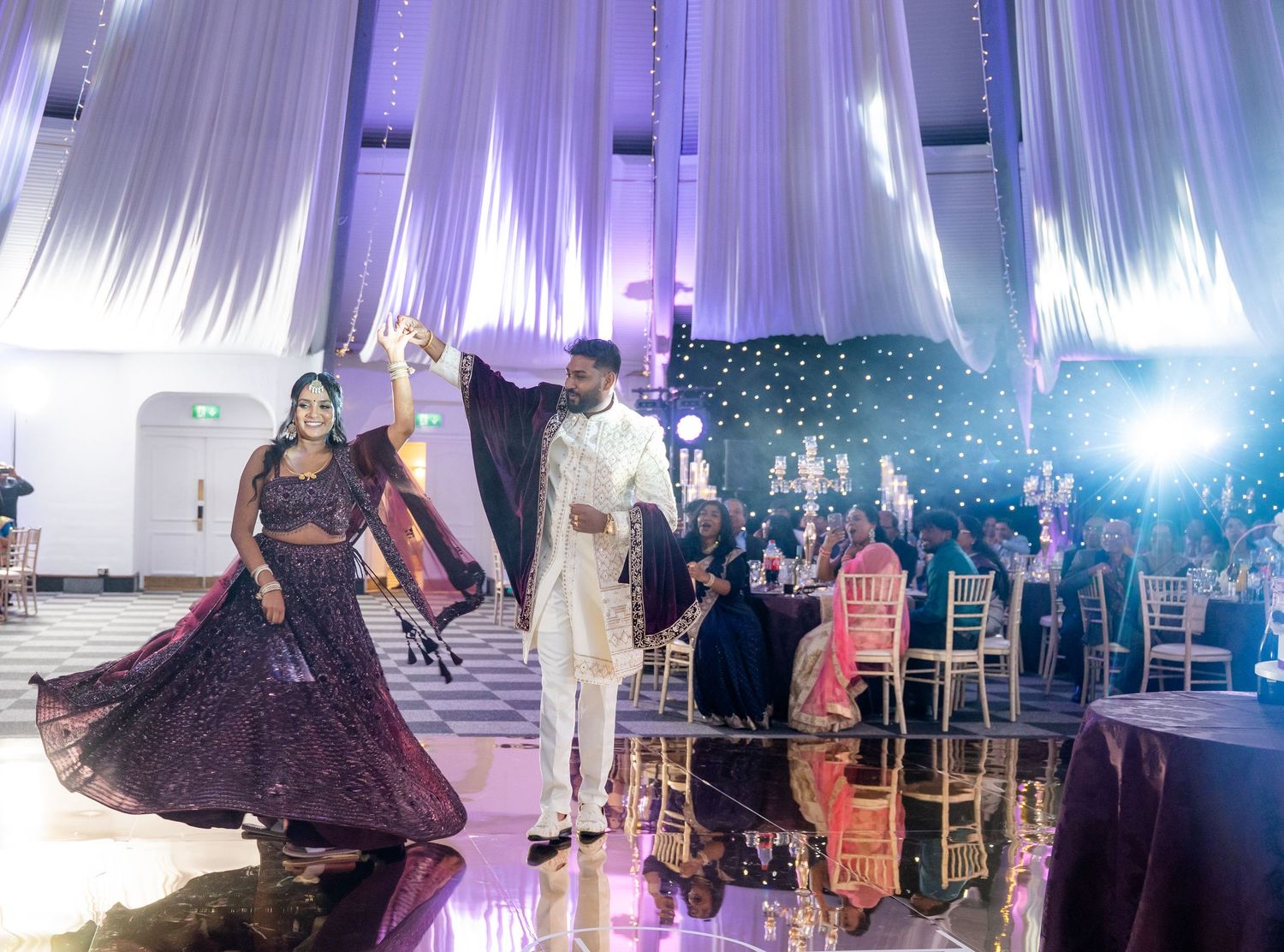 Couple dancing at an elegant Indian wedding reception with purple lighting and white draping.