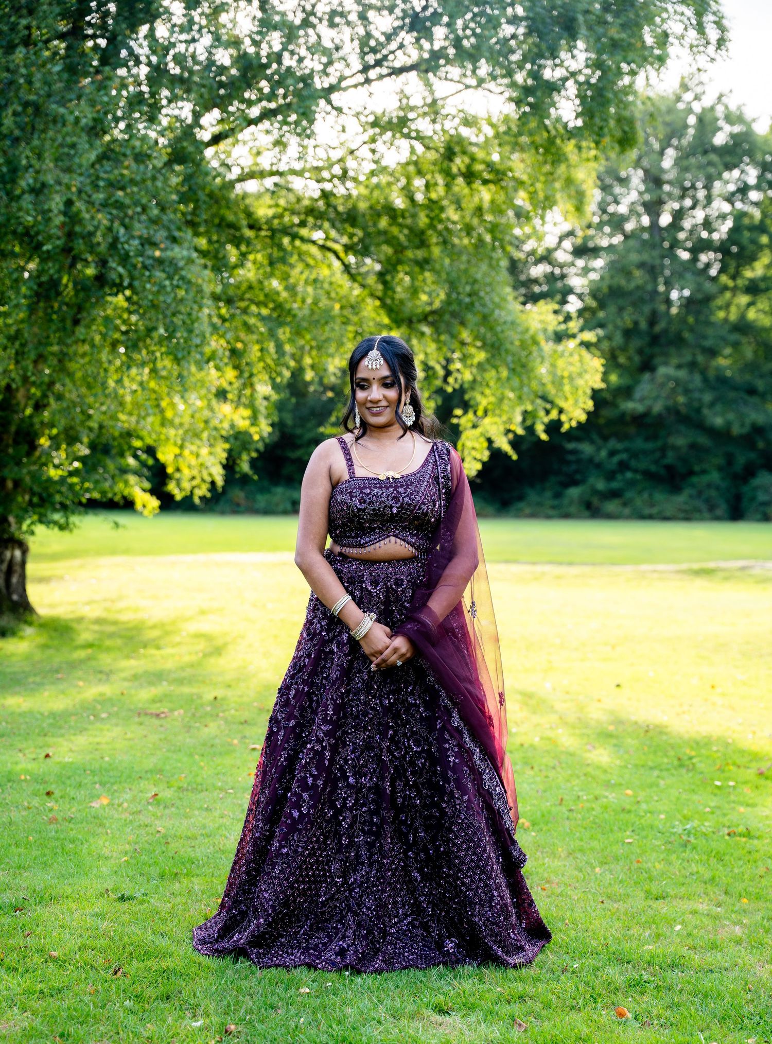 Woman in purple embellished lehenga choli with dupatta standing on green lawn surrounded by lush trees.