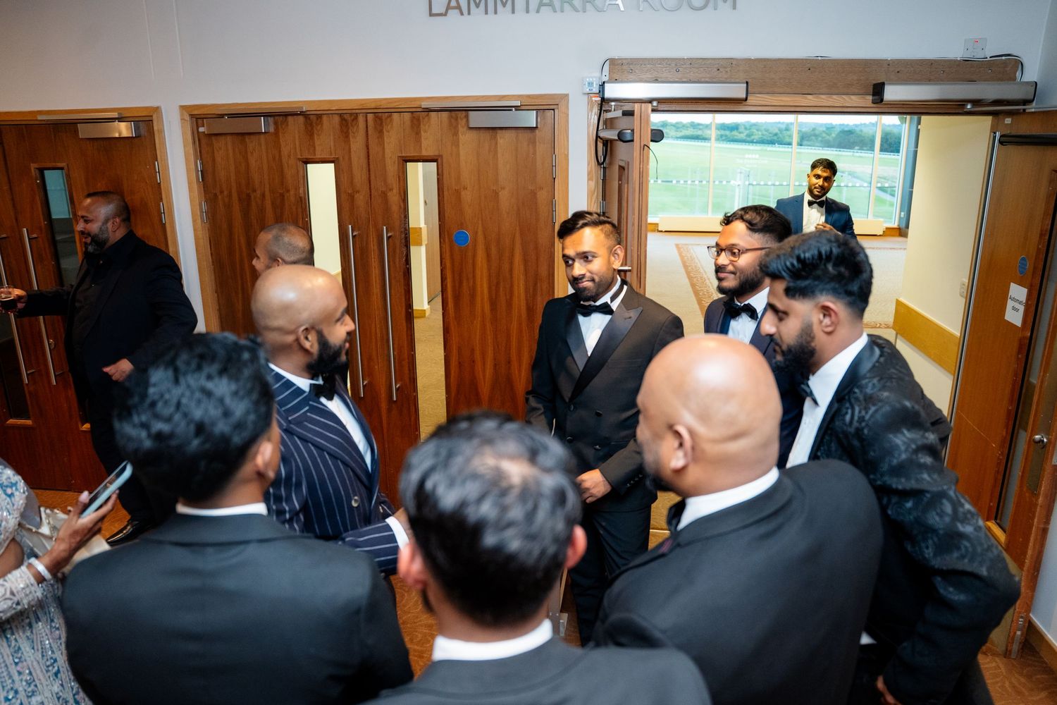 Group of men in formal suits gathered and conversing outside a banquet room door.