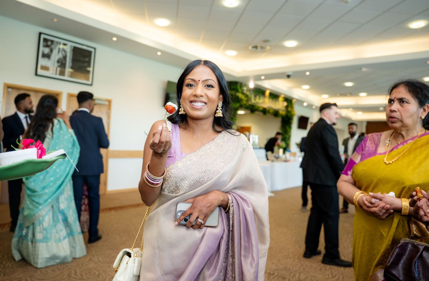 Woman in silver saree smiling and holding food at an Indian wedding reception gathering.
