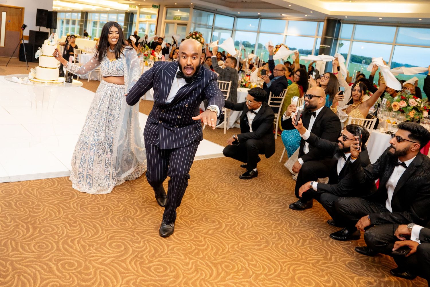 Groom in pinstripe suit performs energetic dance move as bride watches at packed wedding reception.