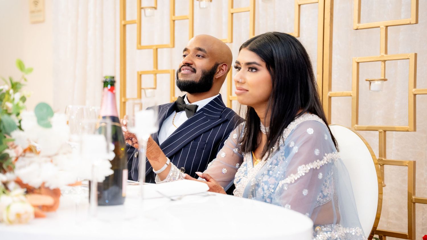 A bride in a sheer blue gown and groom in a pinstripe suit sit attentively at their wedding reception.