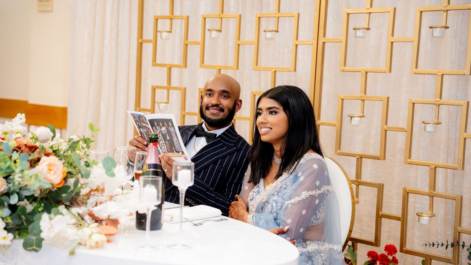 A bride and groom in elegant wedding attire sit at a decorated sweetheart table reviewing a magazine.