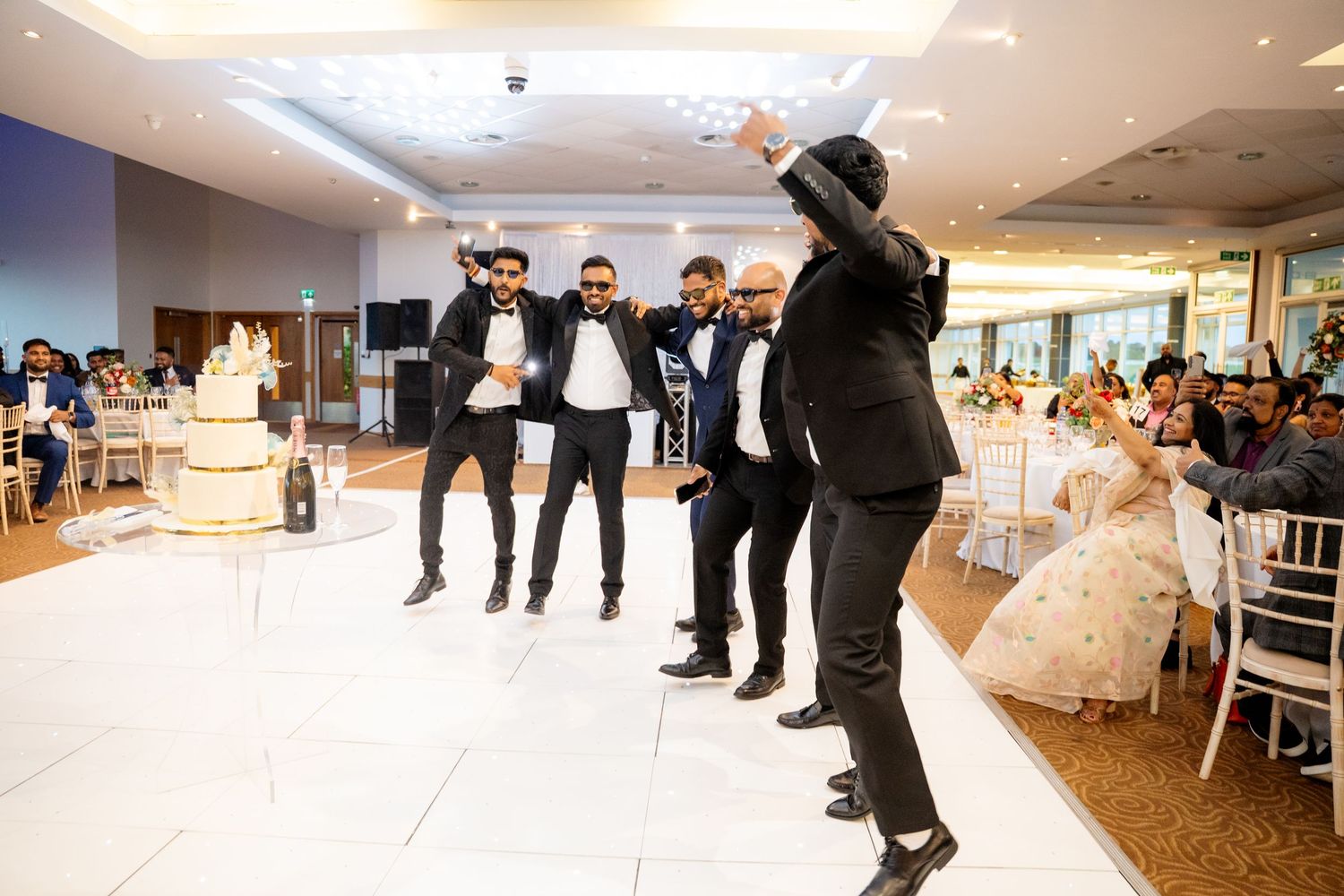 Groomsmen in black tuxedos performing a surprise dance routine near the wedding cake at reception.