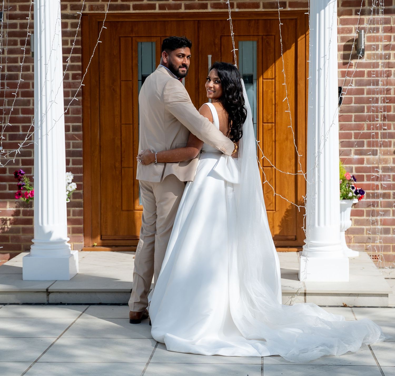 A bride and groom pose together in front of a brick building with white columns on their wedding day.
