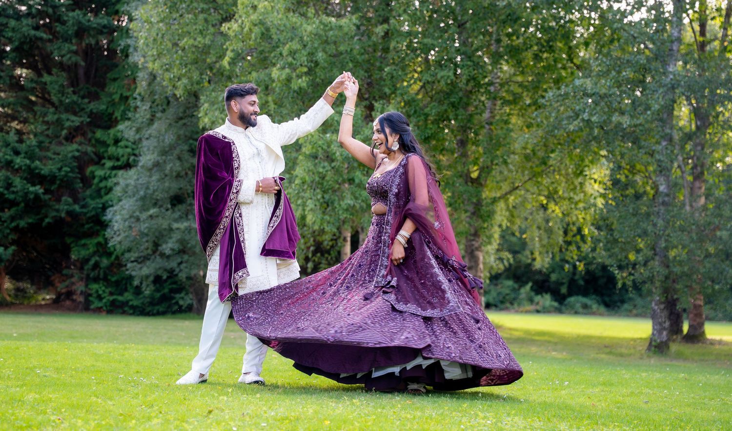 Bride in purple lehenga twirls gracefully while groom spins her in a lush green outdoor setting.