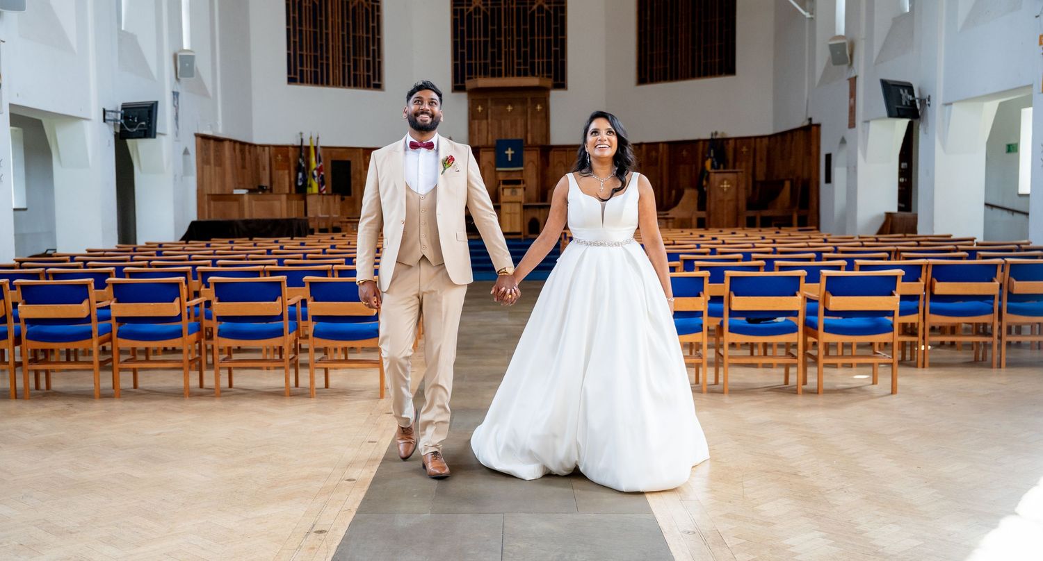 Bride and groom holding hands walking down the aisle of an empty church with blue chairs.