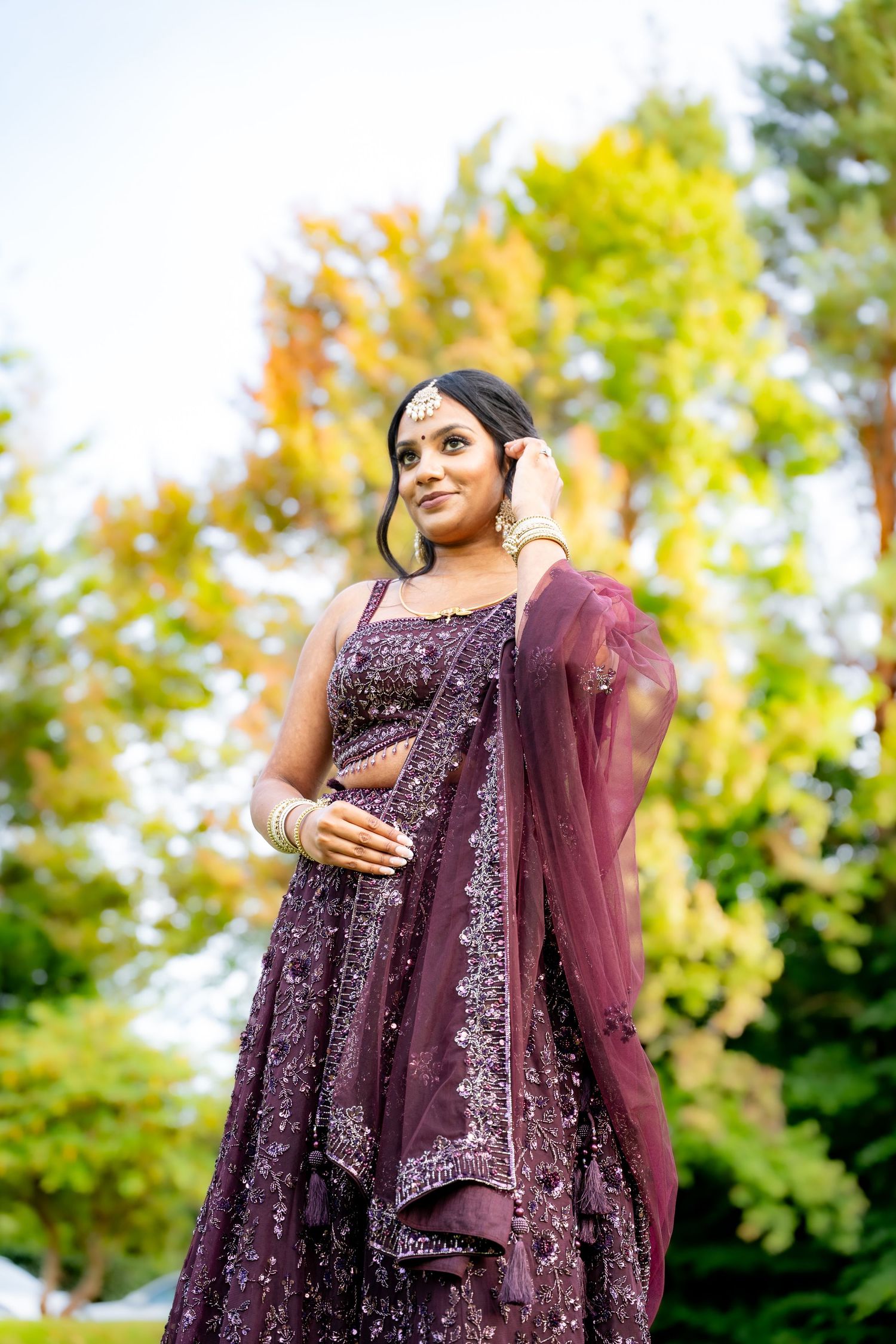 A bride in a purple sequined lehenga with dupatta poses outdoors surrounded by autumn trees.