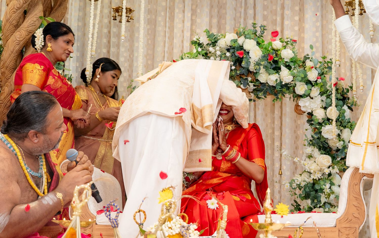 Groom leaning toward seated bride during intimate South Indian wedding ritual with priest chanting blessings.