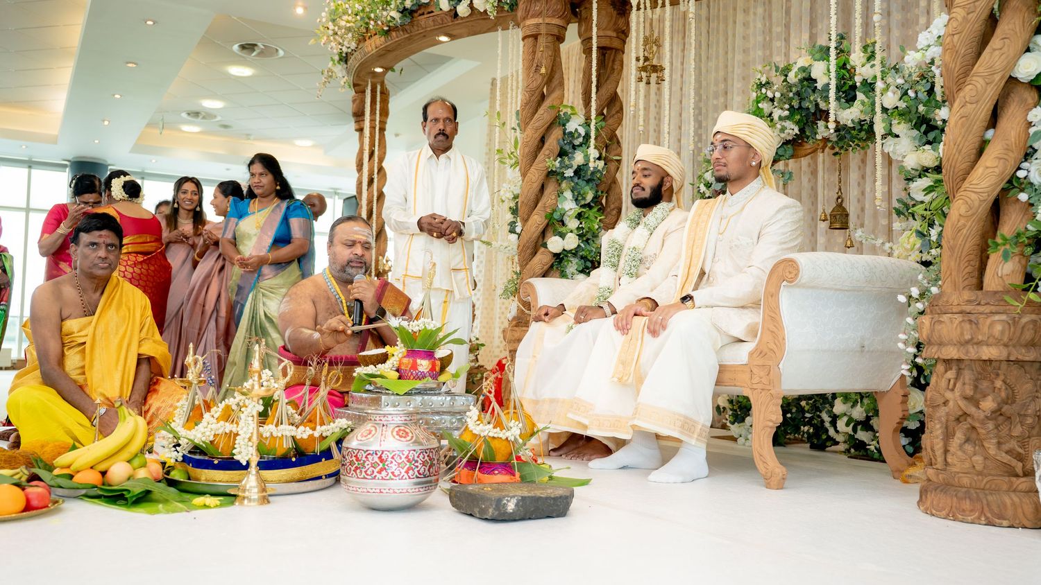 Two grooms in white sit at a Hindu wedding ceremony with priests and guests in colorful attire.