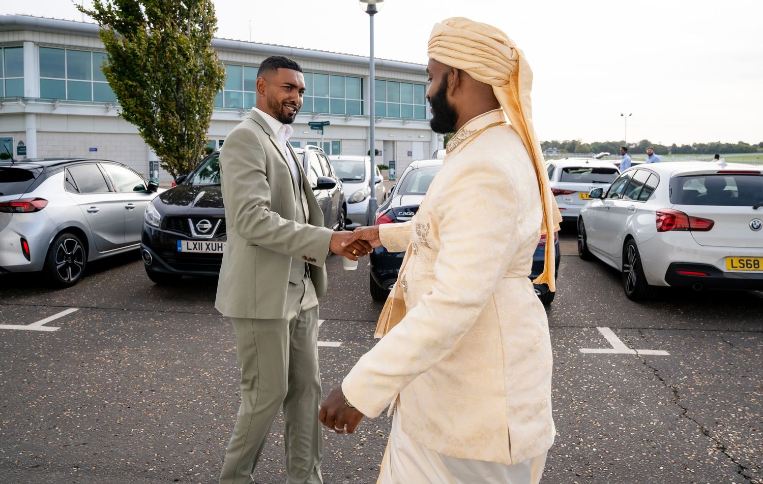 Two men in formal attire shaking hands in a parking lot, one in a suit and one in traditional South Asian dress.