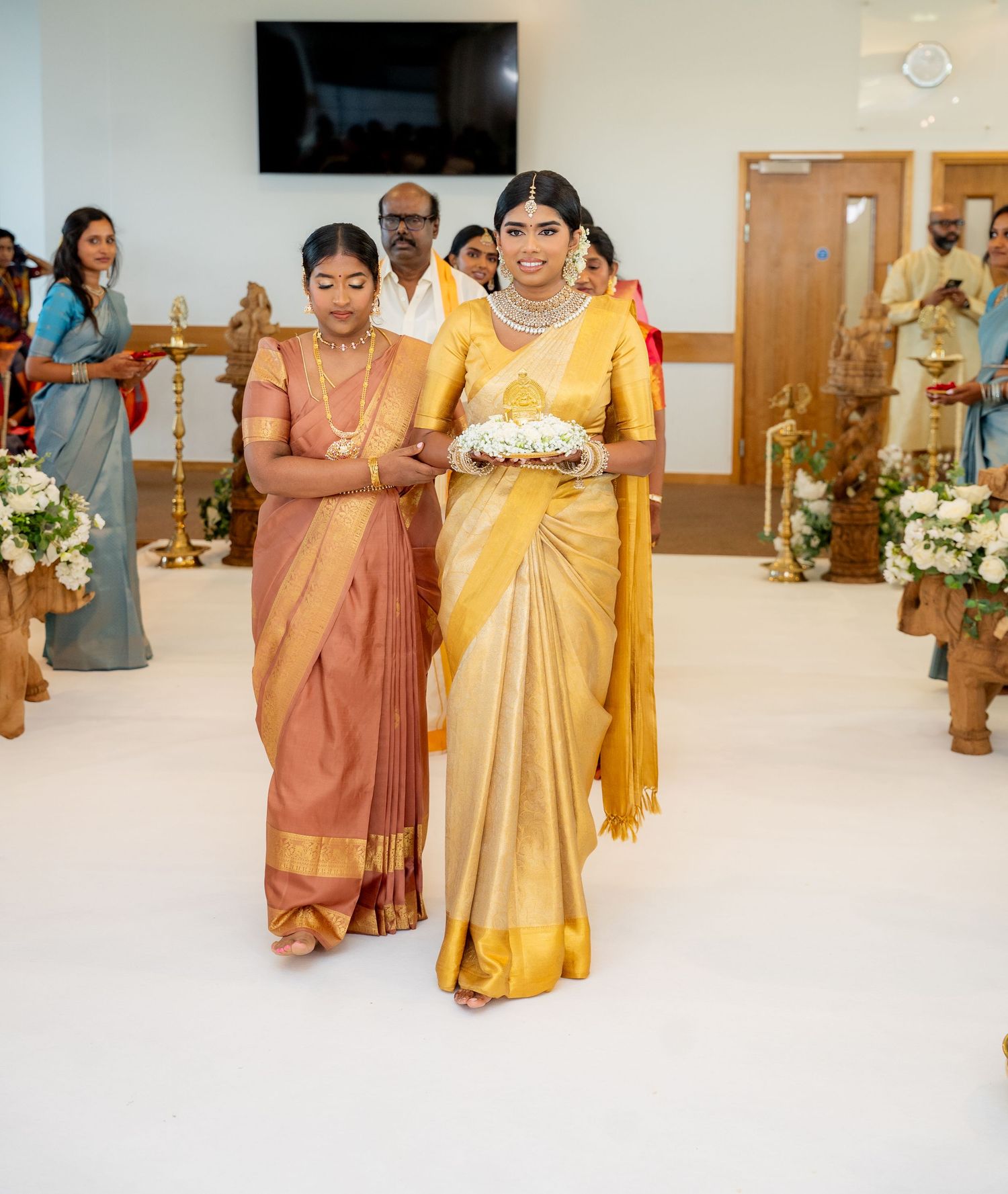 Two women in traditional silk sarees walking together at an indoor ceremony with guests and floral decorations in the background.