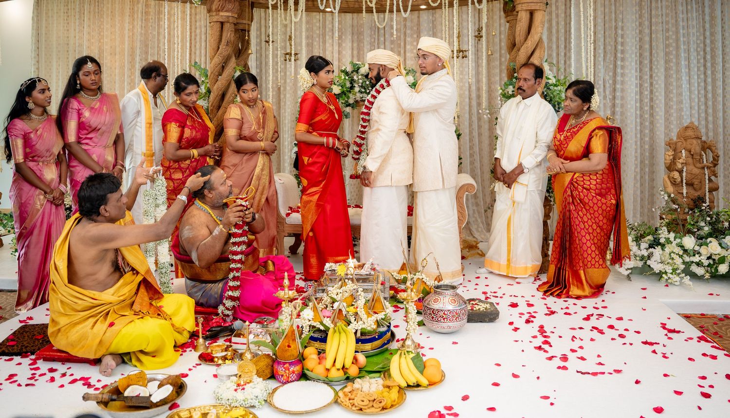 Traditional Hindu wedding ceremony with priest performing rituals as couple stands under floral mandap with guests.