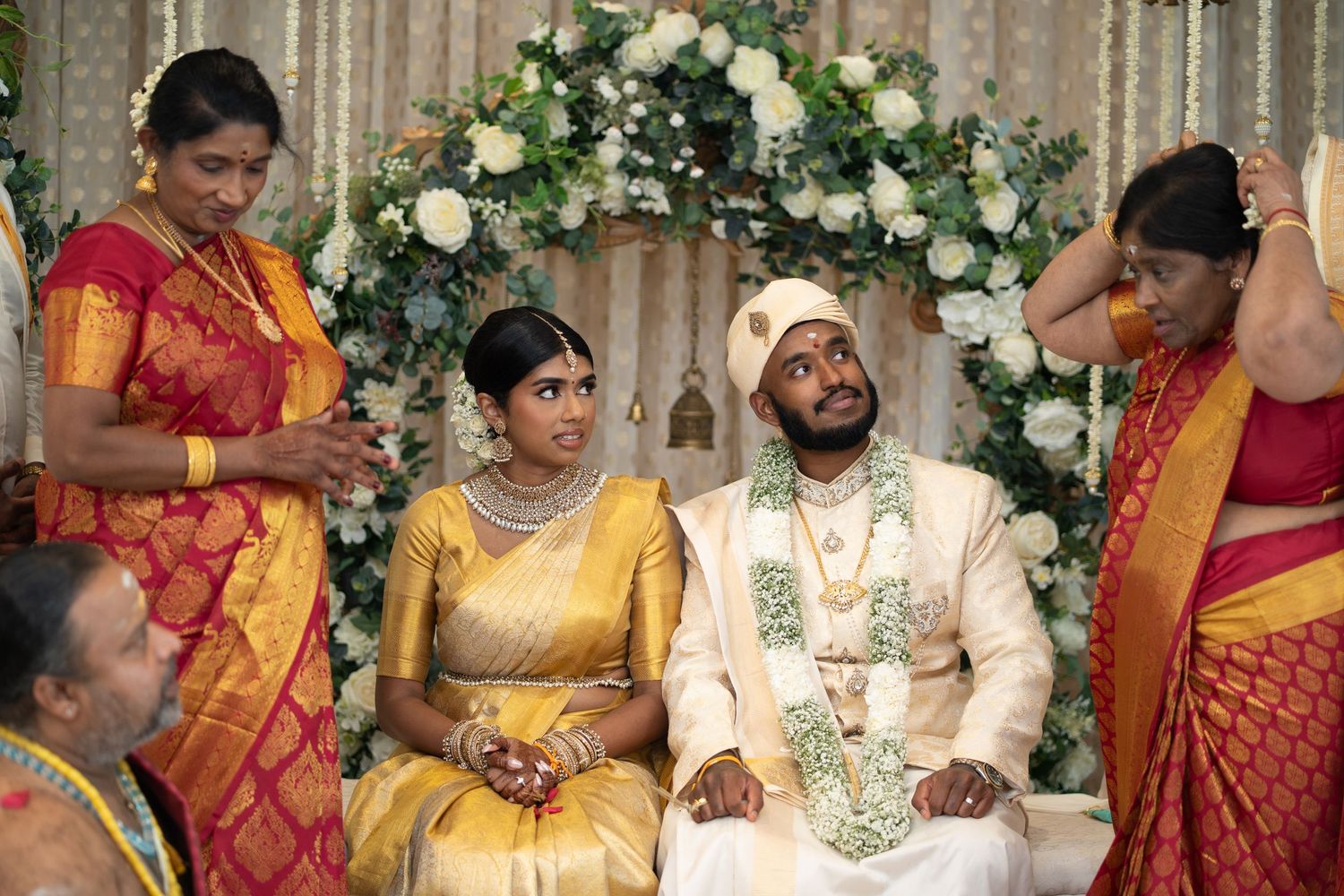 Bride in gold saree and groom in cream sherwani sit together during a traditional South Indian wedding ceremony with family.