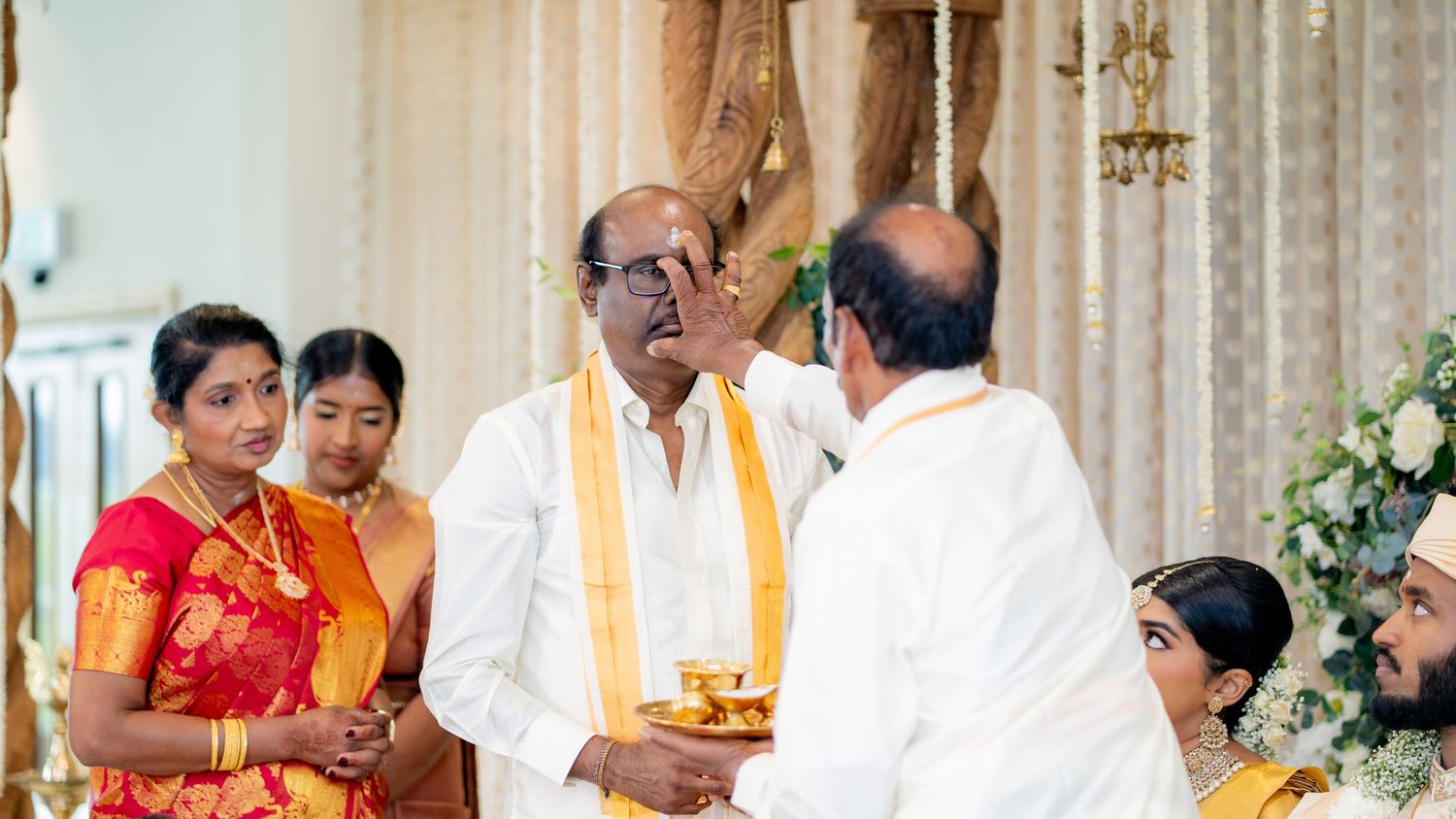 Elderly man receiving traditional blessing with tilak at South Indian Hindu wedding ceremony with family present.