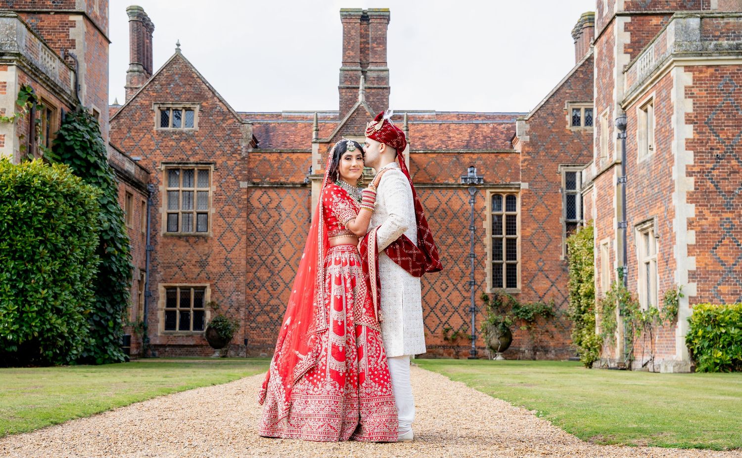 Indian bride and groom in traditional red and white wedding attire posing at a grand brick manor house.