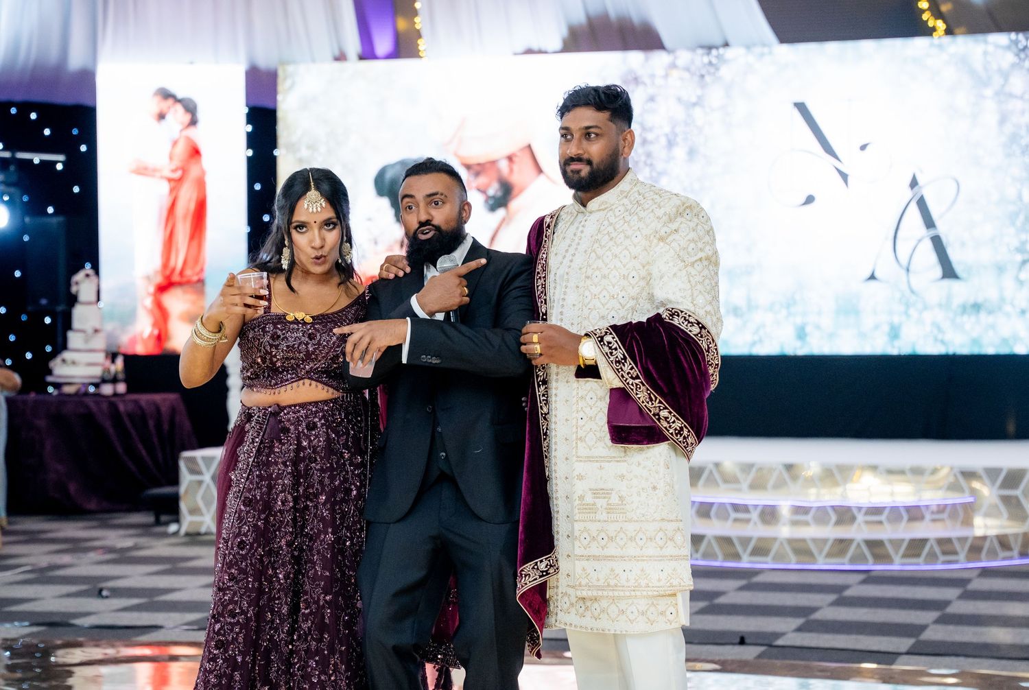 Bride in purple lehenga and groom in sherwani posing with guest at Indian wedding celebration.