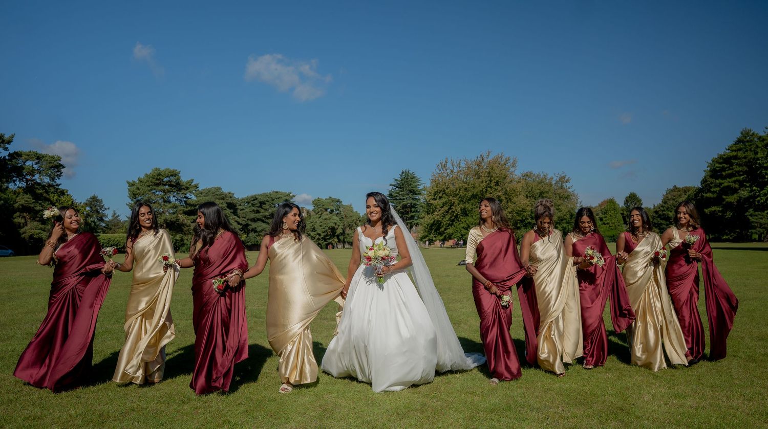 Bride with large bridal party in burgundy and gold gowns walking across green lawn outdoors.