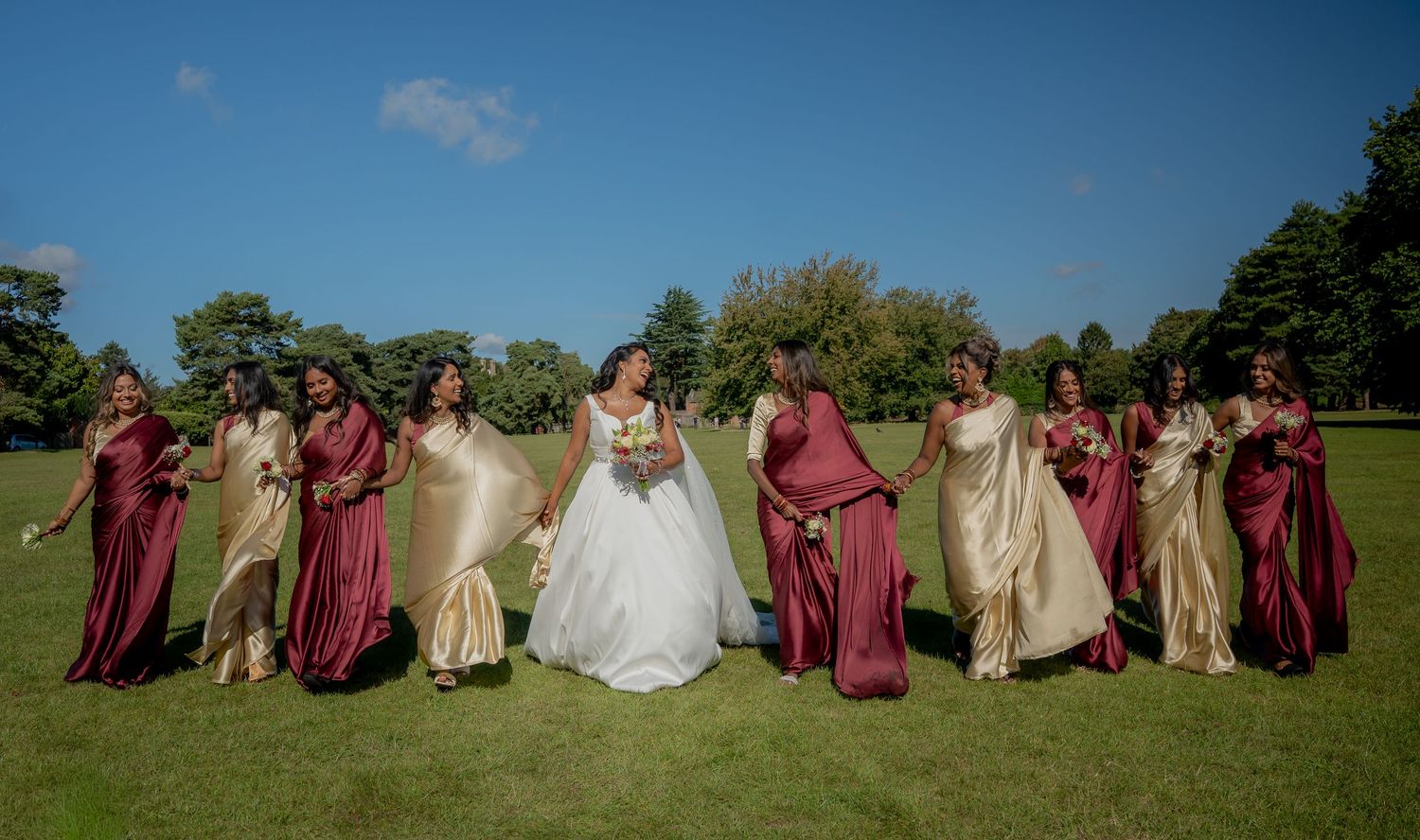 Bride and bridesmaids in burgundy and gold dresses walk playfully across green lawn on sunny wedding day.