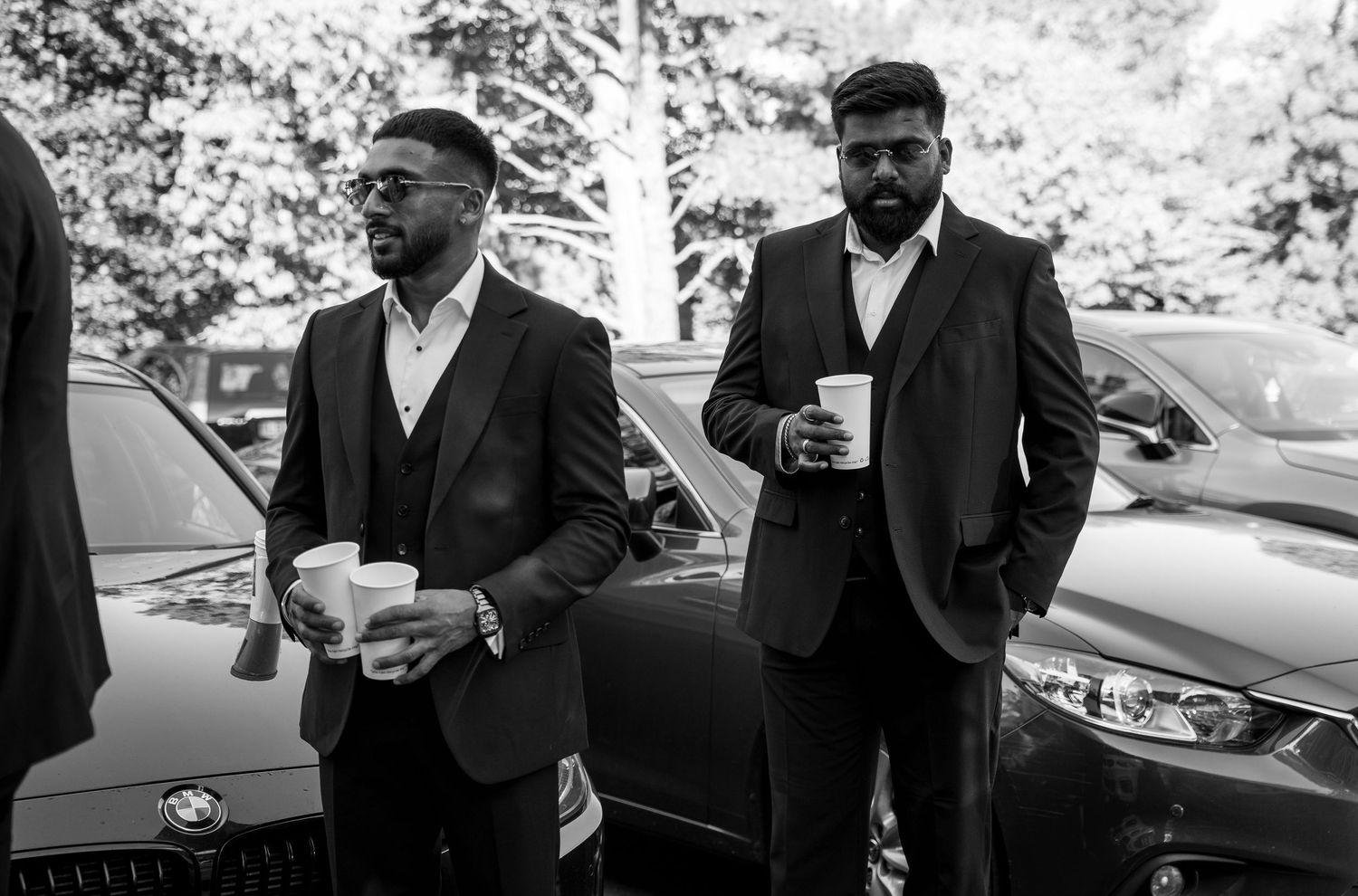 Two men in black suits holding coffee cups stand near luxury cars at an outdoor wedding event.
