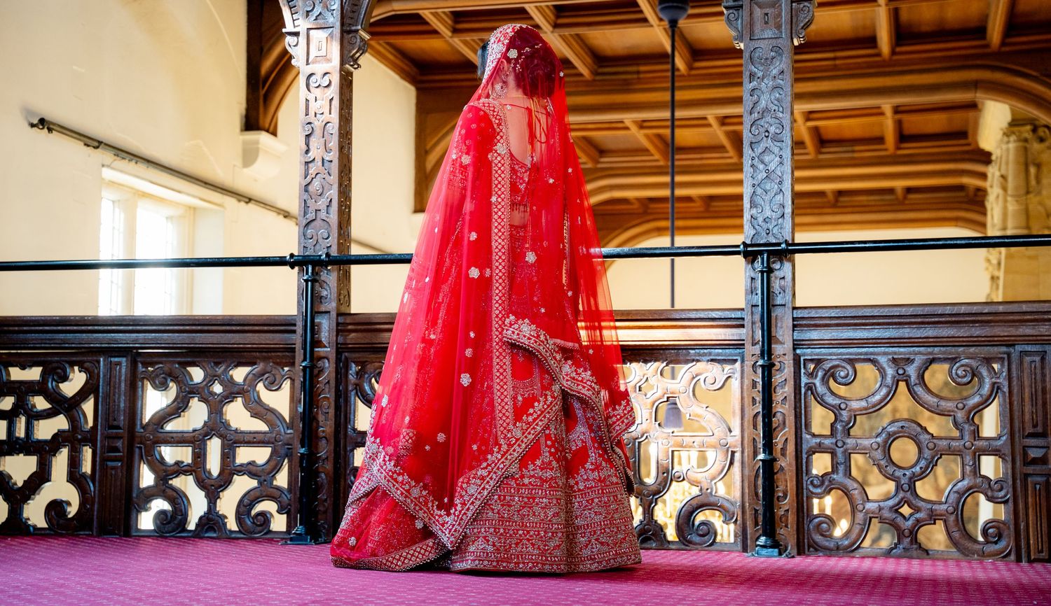 Indian bride in stunning red embroidered lehenga with dupatta veil standing on ornate wooden balcony.