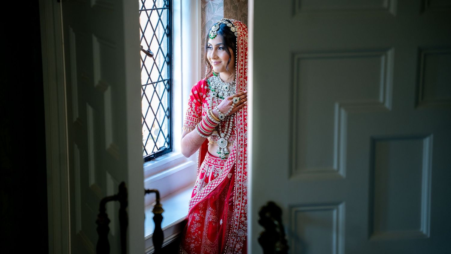 Indian bride in red lehenga stands elegantly by window in soft natural light.