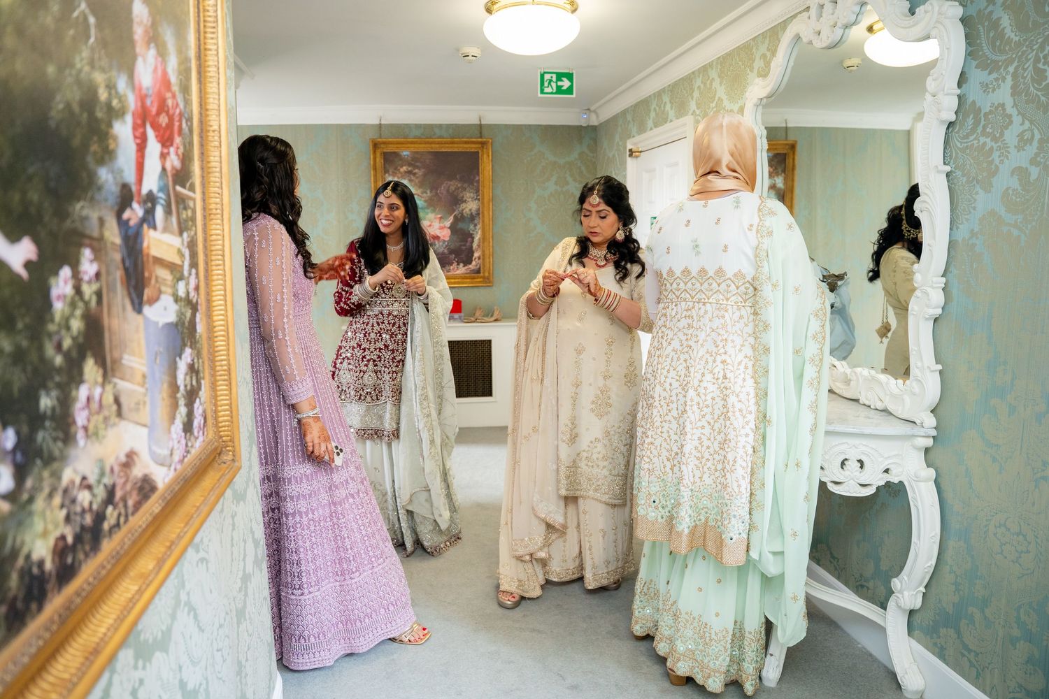 Three women in colorful Indian outfits chat near a mirror during wedding preparations.