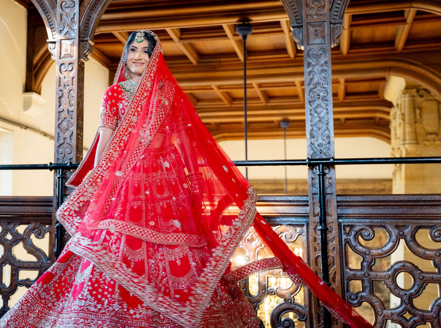 Bride in stunning red bridal lehenga with gold embroidery and dupatta posing on ornate balcony.