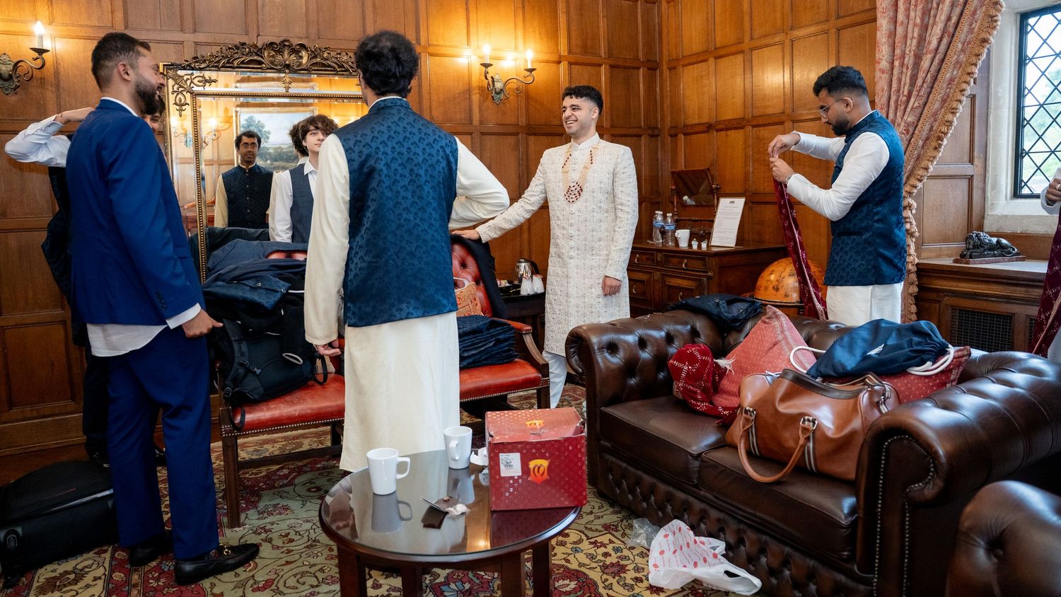 Men in traditional South Asian attire prepare for a wedding ceremony in an elegant room with leather sofas.