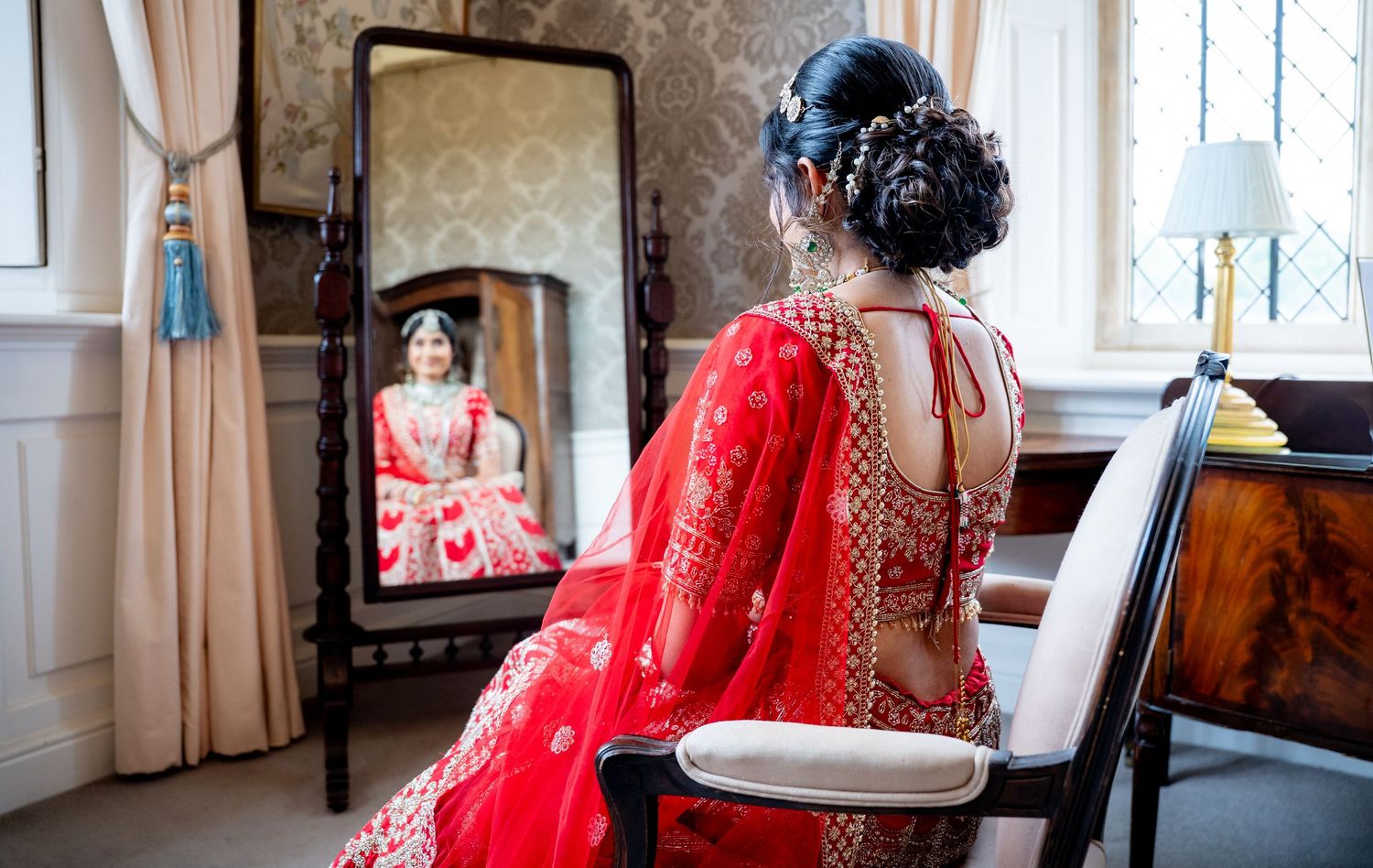Indian bride in red lehenga seated before full-length mirror reflecting her traditional bridal attire.
