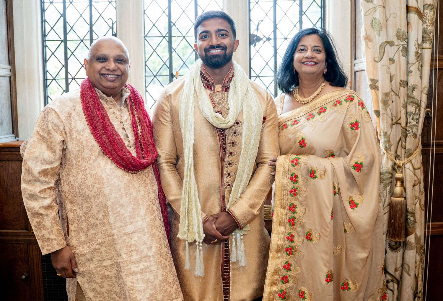 South Asian groom in gold sherwani posing with parents in traditional Indian wedding attire.