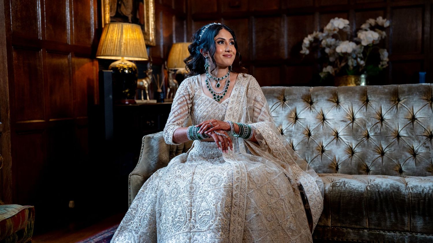 A woman in an elegant white embroidered lehenga sits gracefully on a tufted sofa in a warmly lit wood-paneled room.