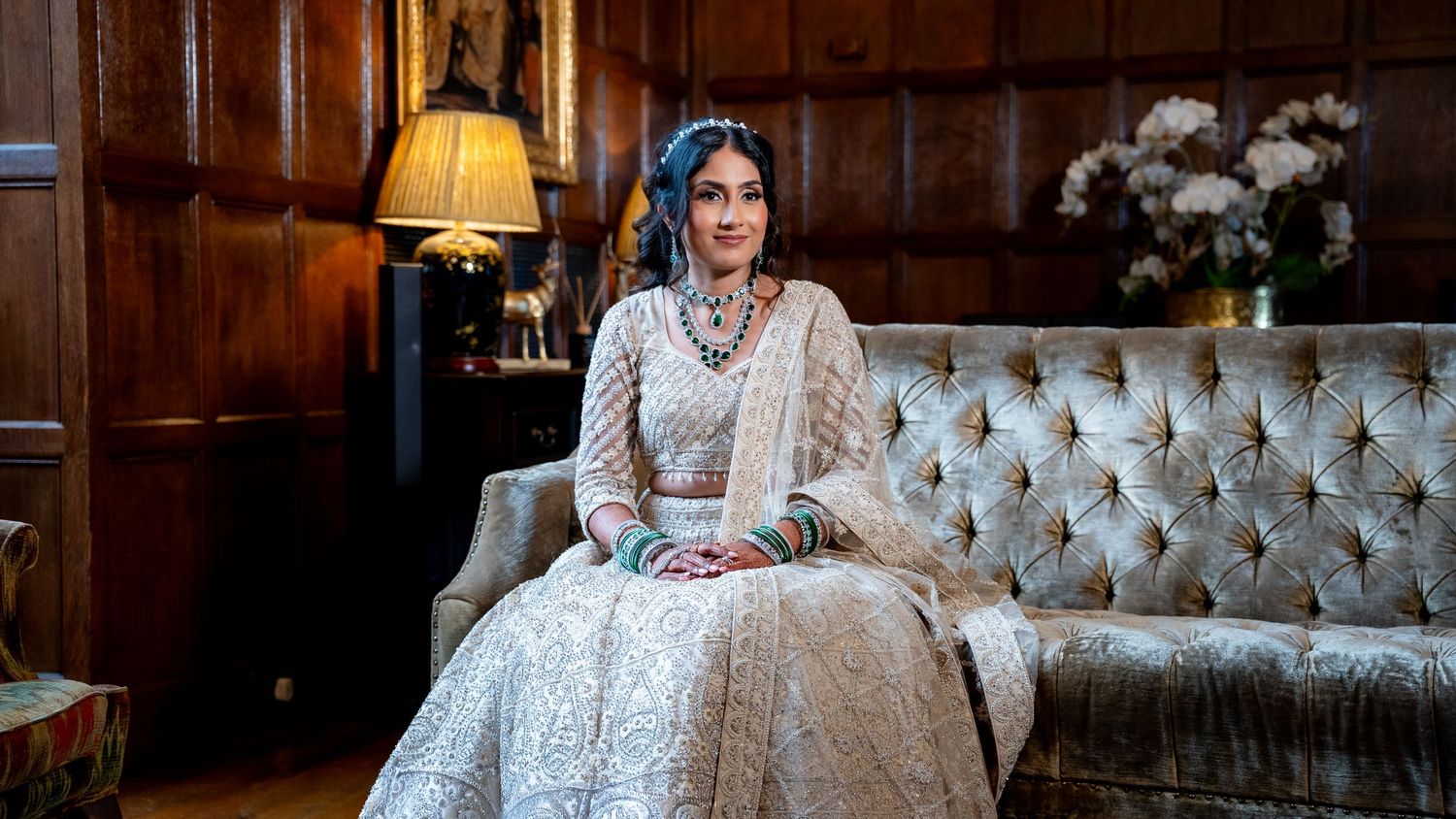 Woman in elegant white embroidered South Asian bridal lehenga seated on tufted sofa in ornate wood-paneled room.