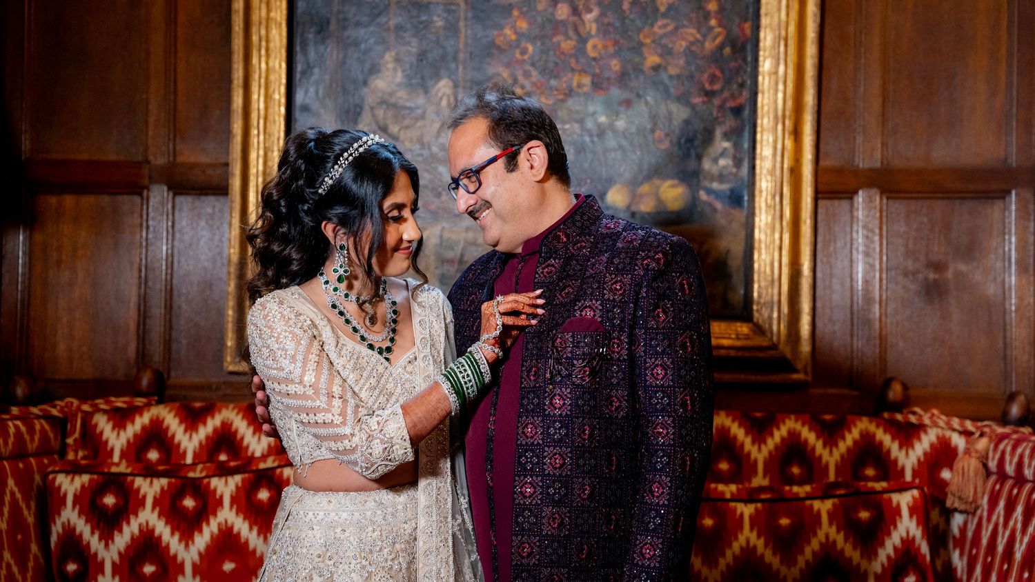 Indian couple in traditional wedding attire smiling at each other in an elegant wood-paneled room.