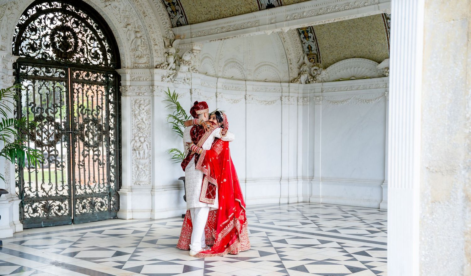 Couple in traditional Indian wedding attire embracing in an ornate white marble pavilion with decorative iron gates.