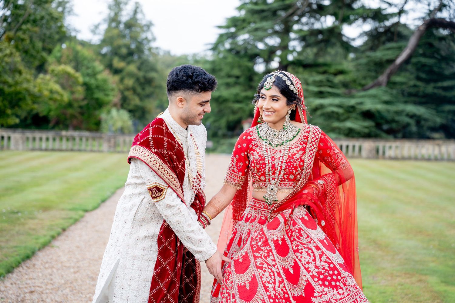 Indian bride in red lehenga and groom in white sherwani hold hands smiling in garden.