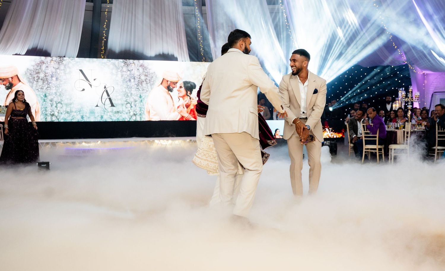 Two grooms share a joyful first dance surrounded by dramatic fog at an elegant wedding reception.