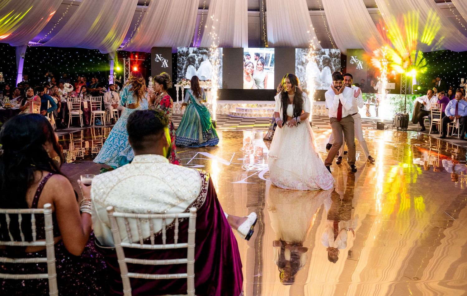 Elegant wedding reception with guests dancing on a reflective floor under a decorated tent with warm lighting.