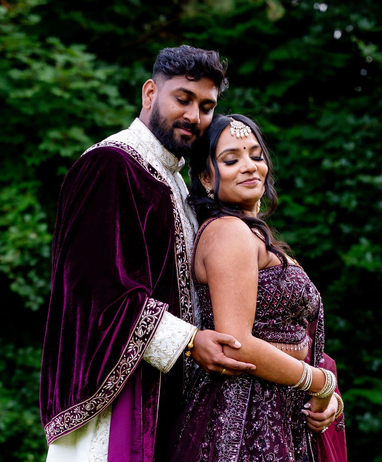 Couple in matching purple traditional Indian wedding attire embracing outdoors with lush green trees in background.